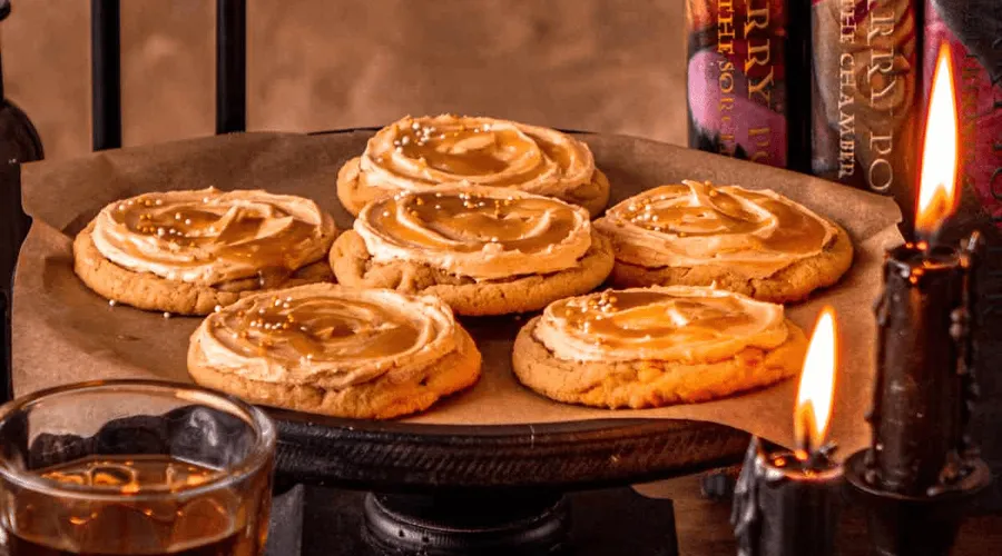 Six frosted butterbeer cookies on a wooden cake stand surrounded by black candles, faux Harry Potter books, and a mug of golden butterbeer — a cozy wizarding-tavern still life.