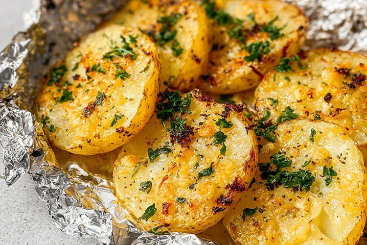 Close-up of golden garlic-butter grilled potato halves in an opened aluminum foil packet, flecked with chopped parsley and cracked pepper.