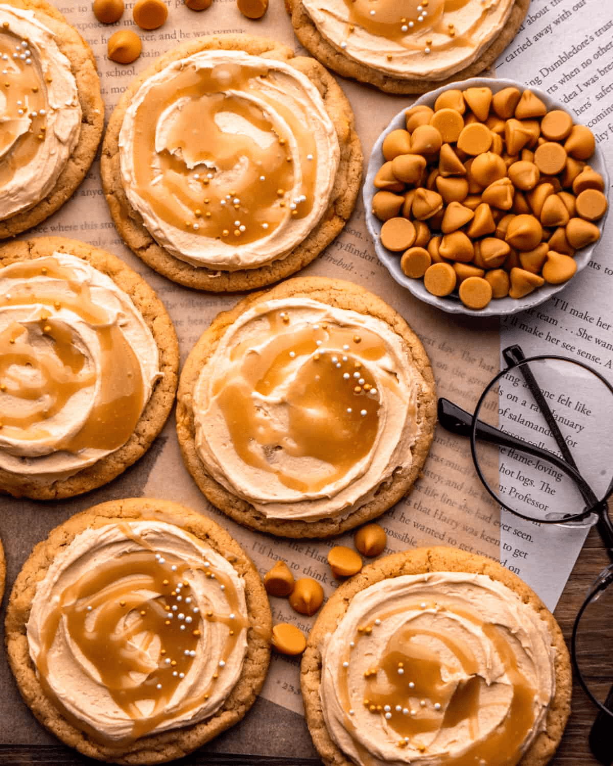 Frosted butterbeer cookies on an open book page — each topped with butterscotch buttercream, drizzled with butterbeer caramel, and dotted with white nonpareils. A bowl of butterscotch chips and round spectacles sit alongside.